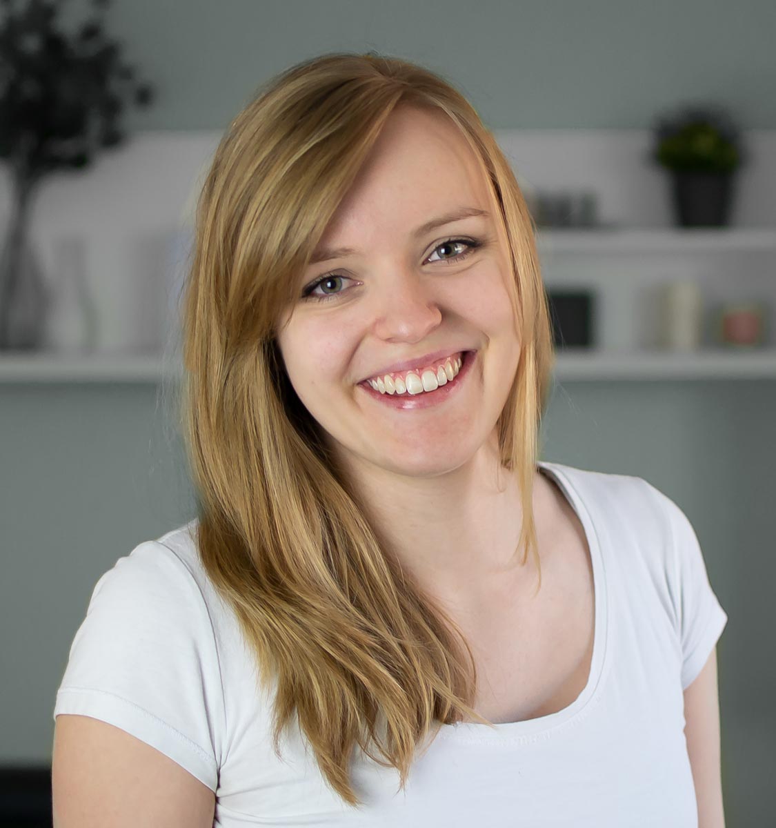 Eine junge Frau mit langen blonden Haaren und einem weißen T-Shirt lächelt in die Kamera. Der Hintergrund ist sanft fokussiert und zeigt Regale mit Dekorationsartikeln und Pflanzen - perfekt für eine Website-Audit- oder Standortanalyse-Fotokulisse.
